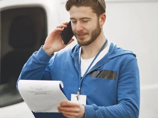 Field worker on a phone call beside a white van, reading a clipboard