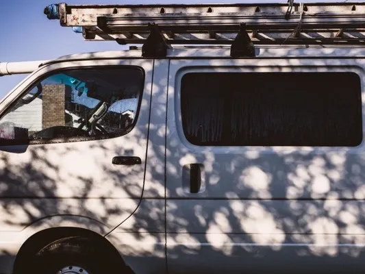 White work van with a ladder on its roof rack, parked in dappled shade
